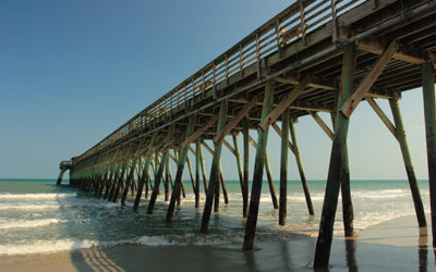 The geometric lines of a fishing pier's posts stand out against the soft hues of green ocean and blue sky.